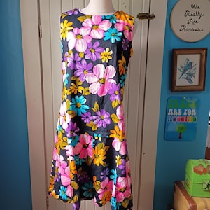 May include: A sleeveless, vintage floral dress on a mannequin. The dress features a black background with large, colorful flowers in shades of pink, purple, yellow, and blue. The dress has a round neckline and a flared skirt.