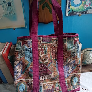 May include: A rectangular tote bag featuring a multi-colored woven tapestry pattern of books, clocks, and writing instruments. The bag has solid dark magenta fabric straps and matching trim along the top and bottom edges, with a structured rectangular shape.
