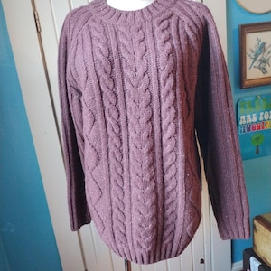 May include: A mauve-colored cable knit sweater displayed on a mannequin. The sweater features a crew neck and long sleeves. The front of the sweater has a prominent cable knit design, with vertical and diamond patterns. The sweater is made of a soft, textured material.