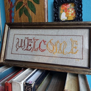 以下が含まれることがあります： 赤い糸、オレンジ色の糸、黄色の糸で「WELCOME」と刺繍されたクロスステッチの額縁。フレームはダークウッド製でシンプルなデザインです。