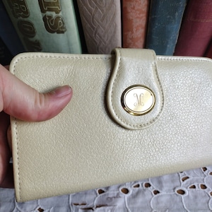 Könnte beinhalten: Eine cremefarbene Ledergeldbörse mit einem goldfarbenen Verschluss und einem Monogramm mit dem Initial "M" auf der Vorderseite.