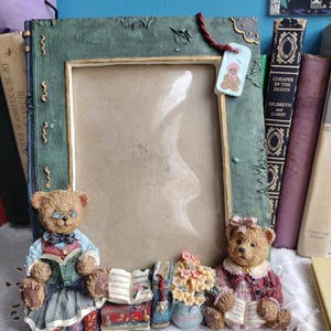 May include: A decorative picture frame with a green, textured border and a gold inner frame. The frame features two teddy bear figurines, one wearing glasses and reading a book, and the other with a pink bow. A small tag with a teddy bear image hangs from the top.