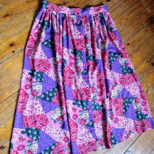 Op de afbeelding: Een vintage patchwork rok in tinten paars, roze en rood. De rok heeft een knoopsluiting en een verscheidenheid aan bloemen- en paisley-patronen. De rok is gemaakt van een lichte stof en heeft een vloeiend silhouet.