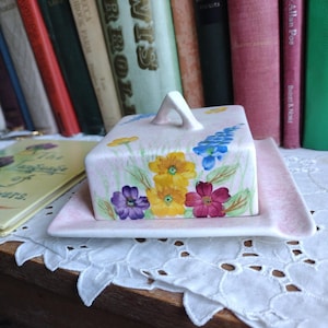 Peut inclure: Un beurrier en céramique rose avec un couvercle et un plateau assorti. Le couvercle est décoré de motifs floraux colorés en jaune, violet et rouge. Le plat est posé devant une collection de livres anciens.