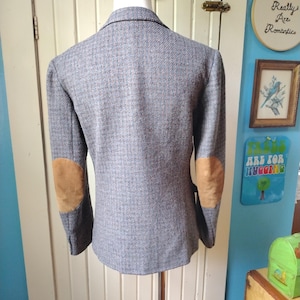 May include: A grey tweed blazer with brown suede elbow patches. The jacket is displayed on a mannequin, highlighting its tailored fit and classic design. The blazer has a two-button closure and notched lapels.