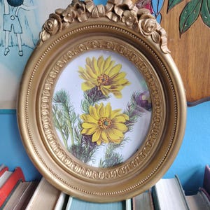 Puede incluir: Un marco de fotos ovalado dorado con un diseño floral. El marco rodea una impresión de flores amarillas con follaje verde. El marco tiene una parte superior decorativa con una cinta y detalles florales. El marco descansa sobre una pila de libros.