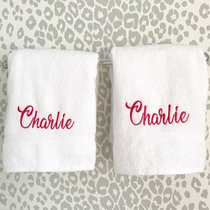 May include: Two white bath towels with the name "Charlie" embroidered in red script. The towels are hanging on a silver bar against a gray and white leopard print wallpaper.