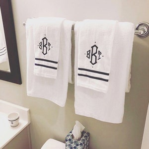 Puede incluir: Dos toallas de ba&ntilde;o blancas con rayas negras y un monograma "BKT" en letra negra colgadas en un toallero cromado.