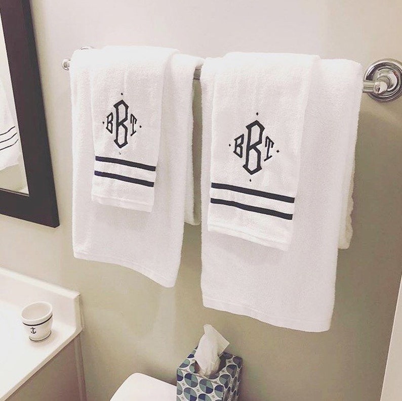Puede incluir: Dos toallas de ba&ntilde;o blancas con rayas negras y un monograma "BKT" en letra negra colgadas en un toallero cromado.