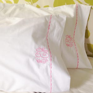 May include: Two white pillowcases with pink embroidered monograms. The pillowcases have a scalloped edge.