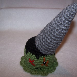 May include: A green and gray crocheted gnome with a pointy hat. The gnome has a stitched-on face with black eyes and a red mouth.
