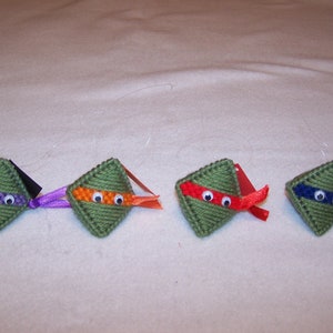 May include: Four green felt ornaments shaped like a turtle head with googly eyes and a colored mask. The masks are purple, orange, red, and blue.