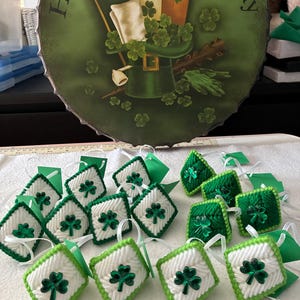 May include: A collection of St. Patrick's Day decorations. The image features green and white diamond-shaped ornaments with shamrock embellishments, along with a round sign that says "Happy St. Patrick's Day" and depicts a leprechaun hat and shamrocks.