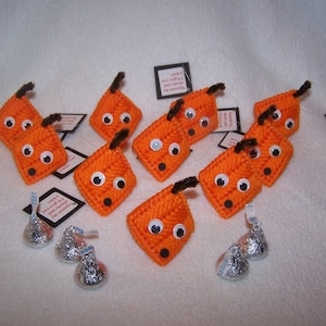 May include: A group of 10 orange felt pumpkins with black yarn stems and googly eyes. The pumpkins are arranged in a semi-circle on a white surface. There are also several silver foil-wrapped chocolate kisses scattered around the pumpkins.