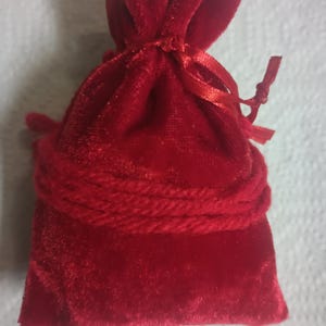 May include: A small, red velvet drawstring bag. The bag is tied at the top with a matching ribbon and a thick, braided cord around the middle. The bag is made of a soft, plush material.