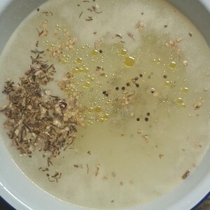 May include: A close-up shot of a white bowl filled with a light yellow liquid. The liquid contains small, dark specks, reddish-brown flakes, and small droplets of oil. The bowl has a blue rim.
