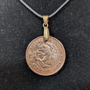 Peut inclure: Pendentif pièce de monnaie cuivrée représentant les armoiries mexicaines, suspendu à un cordon gris foncé. La pièce montre un aigle avec un serpent, entouré de texte. Le pendentif a une bélière dorée.