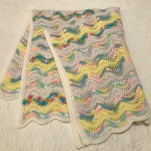 May include: A crocheted baby blanket with a wavy pattern in shades of pink, yellow, blue, and green. The blanket has a white border.