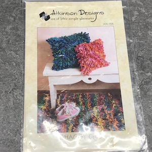 May include: A craft pattern for a shaggy chic rug and cushions. The pattern is called "Shaggy Chic" and the pattern number is ATK-133. The pattern shows a colourful rug and two cushions, one blue and one pink, on a white stool. There are also two pink shoes on the rug.