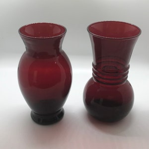 Könnte beinhalten: Zwei rote Glasvasen, eine mit rundem Körper und einem ausgestellten oberen Teil, die andere mit einer traditionelleren Vasenform.