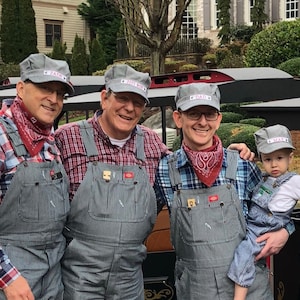 May include: Four men wearing blue and white striped overalls and red and white plaid shirts stand in front of a black train car. They are all wearing matching blue and white baseball caps with the words "City Rail" on them. One man is holding a young boy wearing blue overalls and a white shirt. The train car has a sign that says "Mala Express" on it.