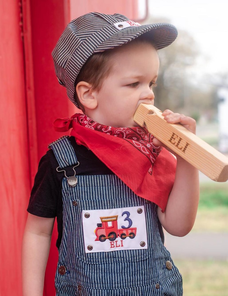 May include: A young child wearing a blue and white striped overall, a red bandana, and a gray and white striped engineer's cap. The child is holding a wooden whistle that says "ELI" on it. The overall has a patch with a red train and the number "3" on it. The patch also says "ELI" on it.