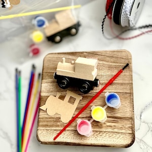 May include: A wooden train craft kit with a wooden train, paintbrushes, paint pots, and ribbon. The kit includes a wooden train cutout and three paint colors: blue, yellow, and pink. The train has black wheels and a natural wood finish.