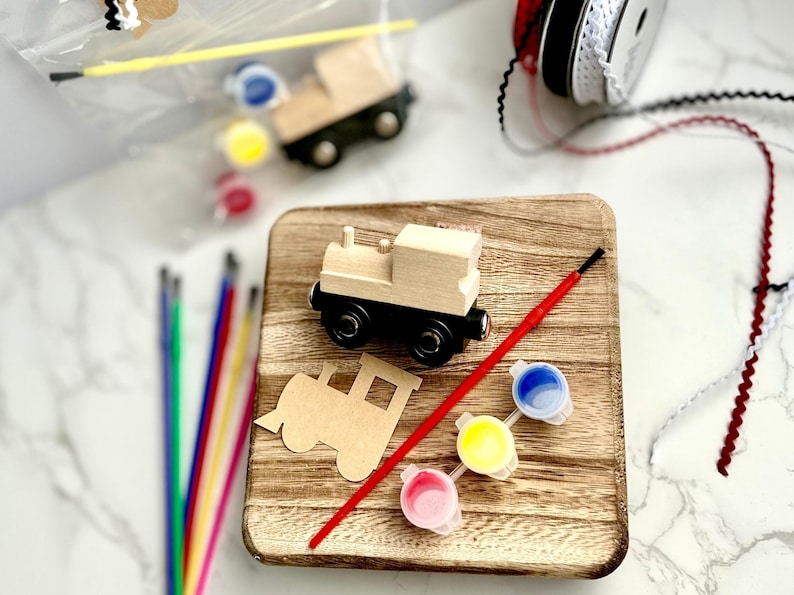 May include: A wooden train craft kit with a wooden train, paintbrushes, paint pots, and ribbon. The kit includes a wooden train cutout and three paint colors: blue, yellow, and pink. The train has black wheels and a natural wood finish.