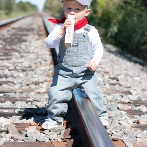 May include: A young child dressed as a train conductor wearing a blue and white striped overalls, a red bandana, and a blue and white striped hat with the name "William" embroidered on it. The child is holding a wooden train whistle and standing on a railroad track.