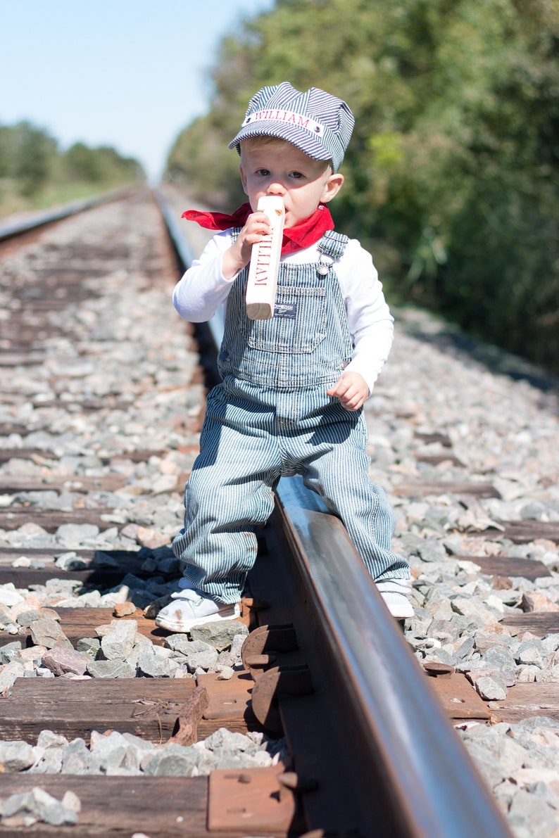 May include: A young child dressed as a train conductor wearing a blue and white striped overalls, a red bandana, and a blue and white striped hat with the name "William" embroidered on it. The child is holding a wooden train whistle and standing on a railroad track.