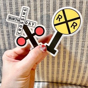 May include: Two stickers: one reads "BIRTHDAY CROSSING" in a railroad crossing sign shape with red circles. The other is a yellow circle with black "R"s and lines, resembling a railroad crossing signal. Both have black and white borders.