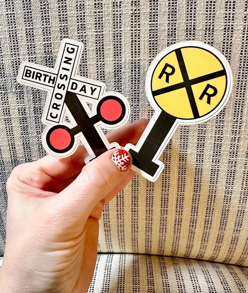 May include: Two stickers: one reads "BIRTHDAY CROSSING" in a railroad crossing sign shape with red circles. The other is a yellow circle with black "R"s and lines, resembling a railroad crossing signal. Both have black and white borders.
