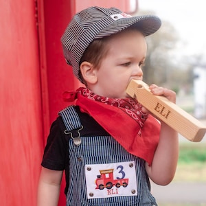 May include: A young child dressed as a train conductor wearing blue and white striped overalls, a red bandana, and a blue and white striped engineer's cap. The child is holding a wooden train whistle that says "ELI" on it. The overalls have a patch with a red train on it that says "ELI" and "3".