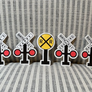 May include: A collection of five railroad-themed birthday decorations. Each decoration features a white background with black and red accents. The decorations include the text "Blake's Day", "Happy Day", "Crossing Day", and "Railroad Day".