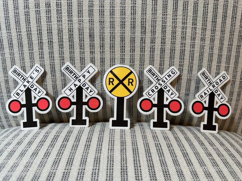 May include: A collection of five railroad-themed birthday decorations. Each decoration features a white background with black and red accents. The decorations include the text "Blake's Day", "Happy Day", "Crossing Day", and "Railroad Day".