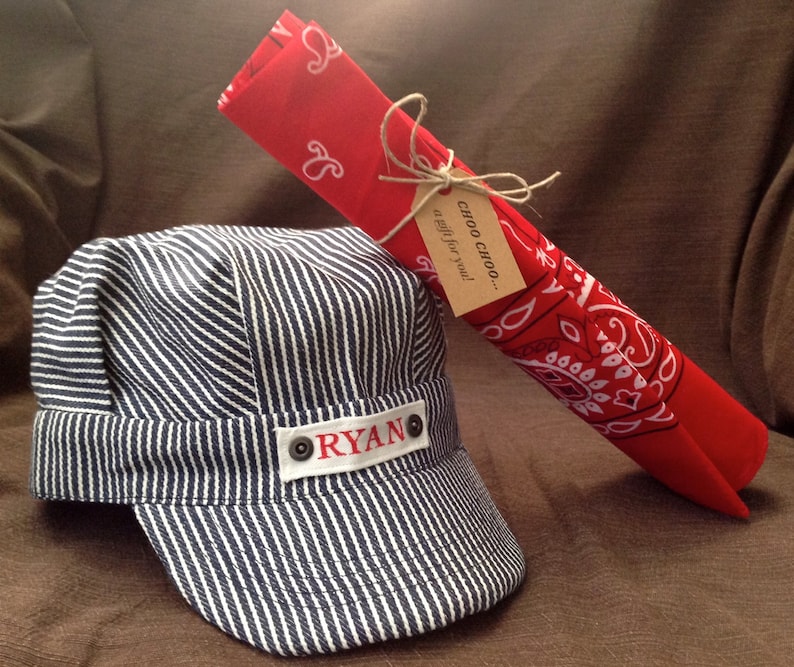 May include: A blue and white striped engineer's cap with the name "RYAN" on the front. A red bandana with a white paisley pattern is tied with twine and a gift tag that reads "Choo Choo... a gift for you!"