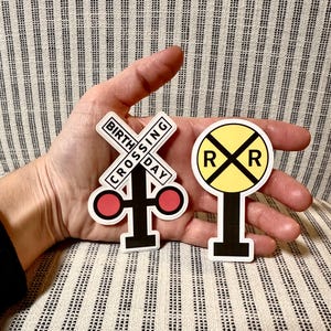 May include: Two stickers featuring railroad crossing signs. One sticker is a white cross with the text "BIRTH CROSSING DAY" and red circles. The other is a yellow circle with an "X" and two "R"s.