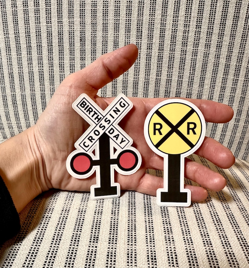 May include: Two stickers featuring railroad crossing signs. One sticker is a white cross with the text "BIRTH CROSSING DAY" and red circles. The other is a yellow circle with an "X" and two "R"s.