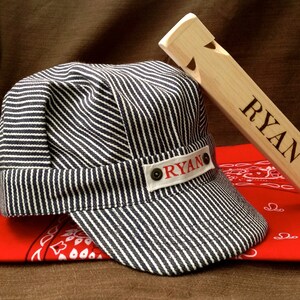 May include: A blue and white striped engineer's cap with a red "RYAN" patch on the side. A red bandana with white paisley print is underneath the cap. A wooden whistle with the name "RYAN" engraved on it is on top of the cap.