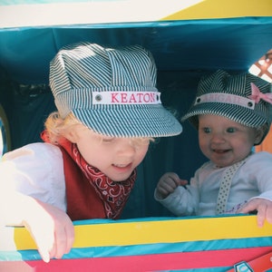 May include: Two children wearing blue and white striped hats with their names, Keaton and Abigail, embroidered on the front. The children are sitting in a colorful play structure.