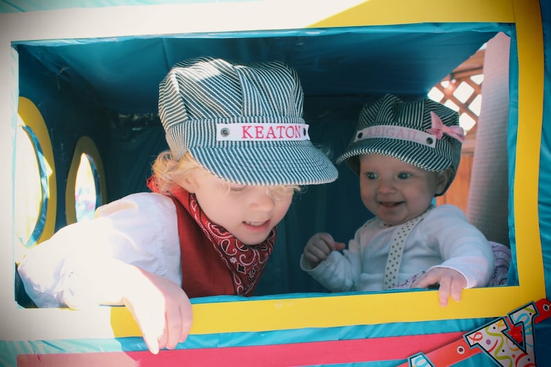 May include: Two children wearing blue and white striped hats with their names, Keaton and Abigail, embroidered on the front. The children are sitting in a colorful play structure.