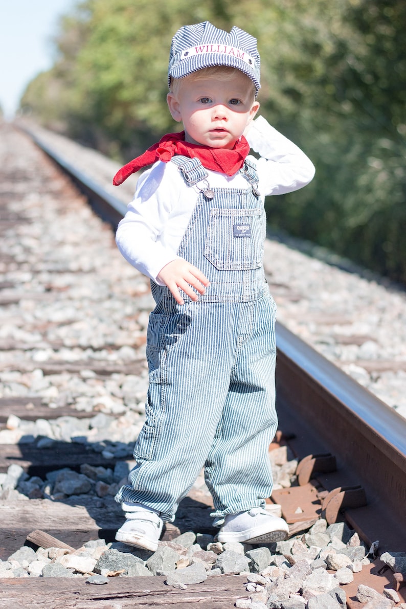 May include: A young child wearing a blue and white striped overalls and a blue and white striped cap with the name "William" embroidered on it. The child is standing on a set of railroad tracks and is wearing a red bandana around their neck.