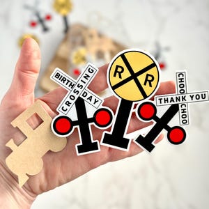 May include: A collection of train-themed stickers and cutouts. Includes a yellow and black railroad crossing sign, crossing signs with red circles, and a train-shaped cutout. Text on the stickers reads "BIRTHDAY CROSSING", "THANK YOU", and "CHOO CHOO".