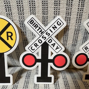 May include: A sticker featuring a black and white railroad crossing sign with the words "BIRTHDAY CROSSING" in a rectangular border. The sign has two red circles on either side. A yellow and black railroad crossing sign is also visible.