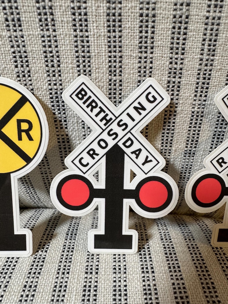 May include: A sticker featuring a black and white railroad crossing sign with the words "BIRTHDAY CROSSING" in a rectangular border. The sign has two red circles on either side. A yellow and black railroad crossing sign is also visible.