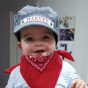 May include: A baby wearing a blue and white striped hat with the name "HARVEY" embroidered on it. The baby is also wearing a red bandana with a white paisley pattern.