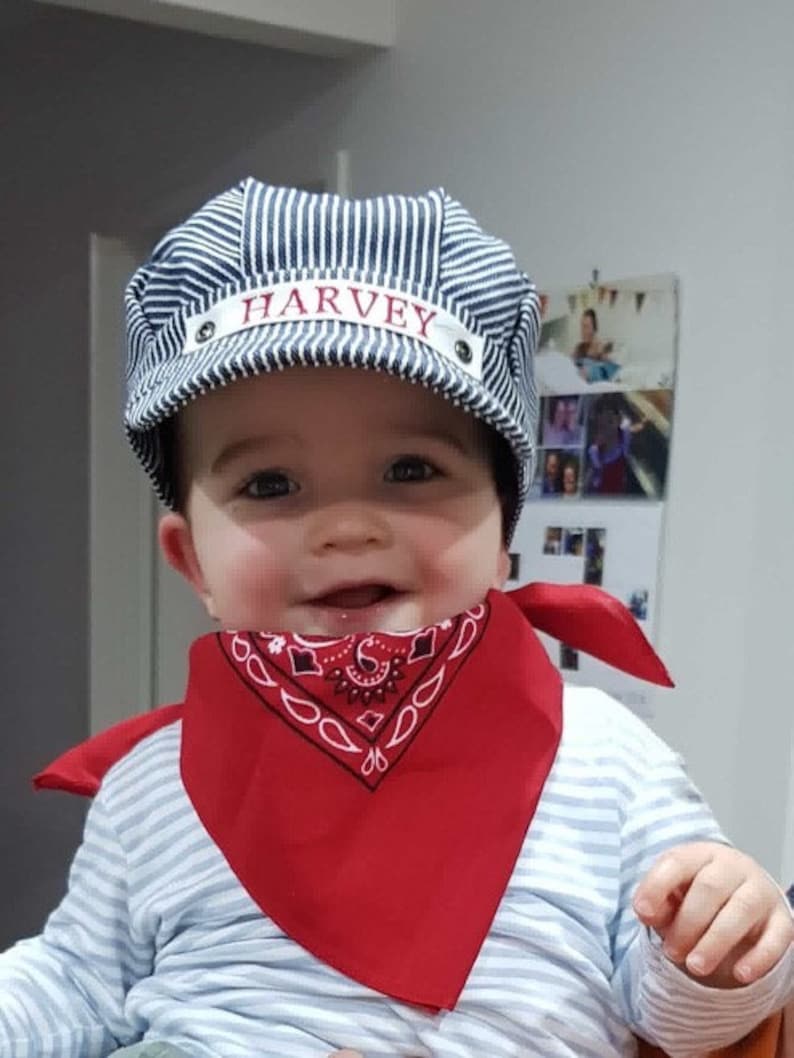 May include: A baby wearing a blue and white striped hat with the name "HARVEY" embroidered on it. The baby is also wearing a red bandana with a white paisley pattern.