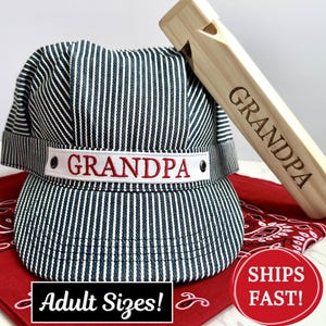 Puede incluir: Gorra de ingeniero a rayas con "GRANDPA" bordado en la parte delantera, junto con un silbato de madera grabado con "GRANDPA". El sombrero es de rayas blancas y negras, y el silbato es de color marrón claro. El sombrero está sobre un pañuelo rojo.