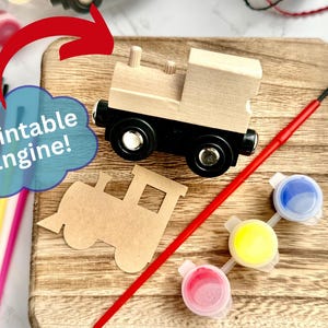 May include: A wooden toy train engine with black wheels and a red paintbrush sits on a wooden board. The image includes three small paint pots with blue, yellow, and pink paint. The text "Paintable Engine!" is in a blue cloud.