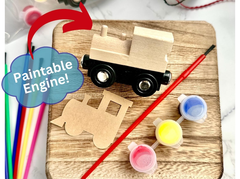 May include: A wooden toy train engine with black wheels and a red paintbrush sits on a wooden board. The image includes three small paint pots with blue, yellow, and pink paint. The text "Paintable Engine!" is in a blue cloud.